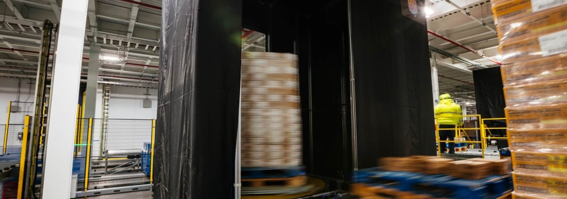 A neatly stacked pallet positioned on a Lineage Eye conveyor system in a clean, well-organized, automated cold storage warehouse, with safety guardrails and scanning stations in the vicinity.